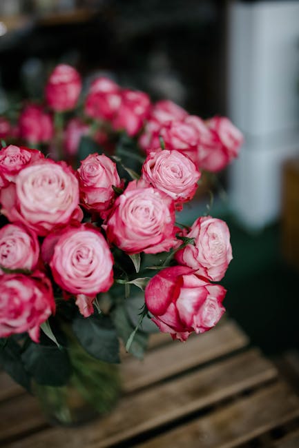 A lush bouquet of pink roses with tightly packed, ruffled petals and glossy green leaves, arranged in a clear glass vase. The bouquet is placed on a wooden surface with a blurred indoor background. Th
