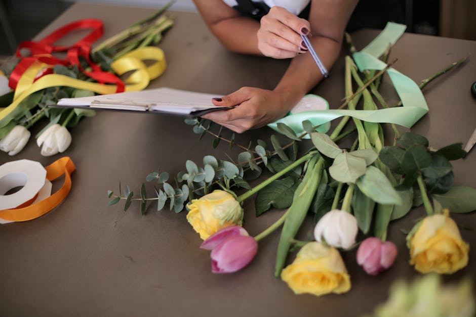 A florist's workspace featuring a variety of fresh flowers including yellow, pink, and white tulips arranged in a loose bouquet, with some stems and leaves visible. The florist's hands are seen holdin