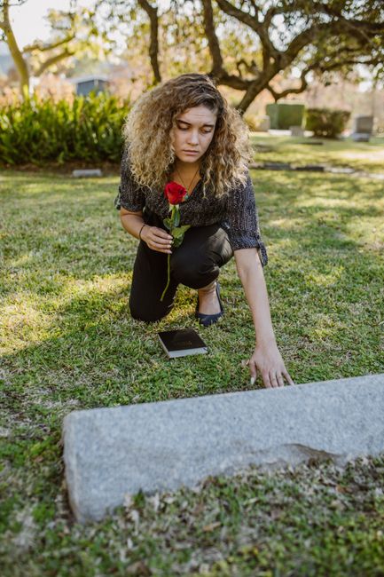 A young woman with curly blonde hair is kneeling on a grassy area in a cemetery, holding a single red rose in her right hand while reaching out with her left hand towards a gray headstone. She is wear