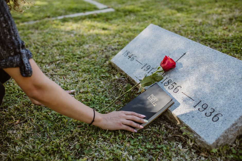 A person is kneeling at a gravestone in a cemetery, holding a small black notebook titled 'Holy Bible' in their left hand while placing a red rose with green leaves on the stone. The gravestone is lig