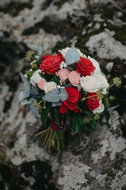A bouquet of fresh flowers featuring red roses, white ranunculus, and blush pink ranunculus, arranged with silvery green eucalyptus leaves and small white filler flowers. The bouquet is expertly wrapp