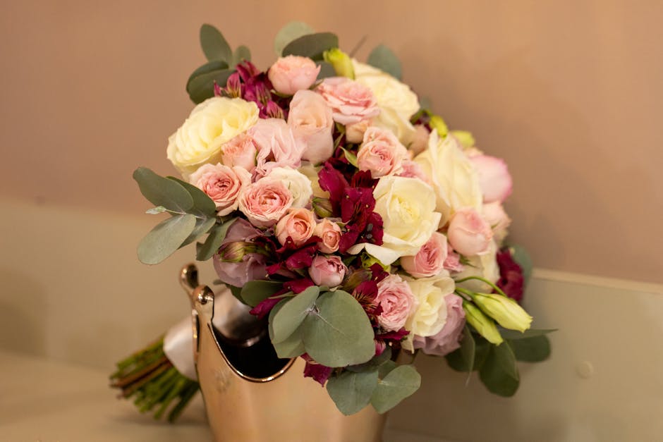 A fresh floral bouquet featuring pale pink and cream roses, complemented by deep burgundy flowers and lush green eucalyptus leaves, arranged in a compact and rounded style. The bouquet is placed in a 