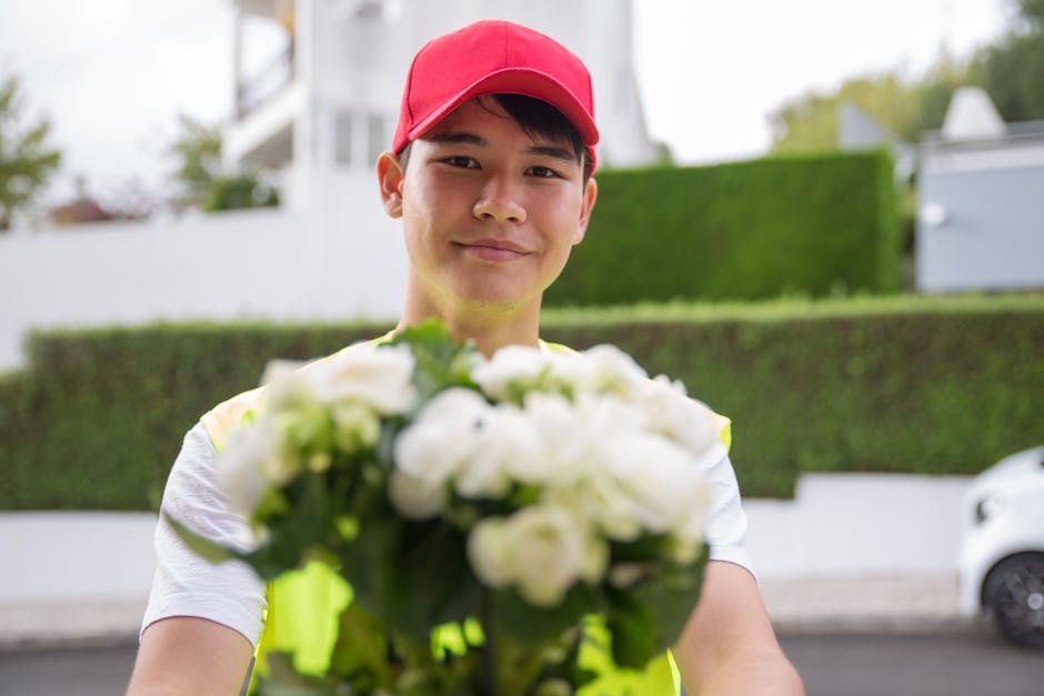 A young delivery person wearing a red cap and a high-visibility vest stands outdoors on a street, smiling as they hold a vibrant bouquet of white roses and green foliage. The bouquet is wrapped in lig